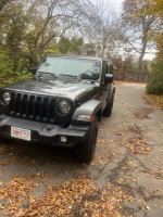 2023 Jeep Wrangler Sport 4WD parked on a leaf-covered driveway, showcasing its grey exterior and rugged design.