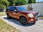 2010 Ford Explorer XLT in orange color parked on a driveway, featuring alloy wheels and tinted windows.