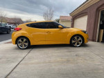 Side view of a yellow 2012 Hyundai Veloster parked in driveway, showcasing its sporty design and alloy wheels.